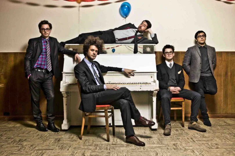 Music band with a white piano and five members in a studio setting.
